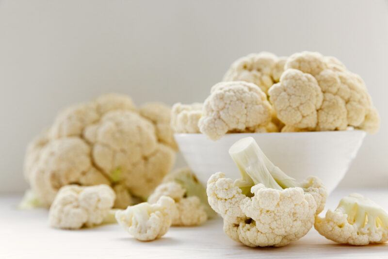 on a white surface is a white bowl full of cauliflower florets, with loose cauliflower florets can be seen in the foreground, while a big cauliflower head can be seen at the back