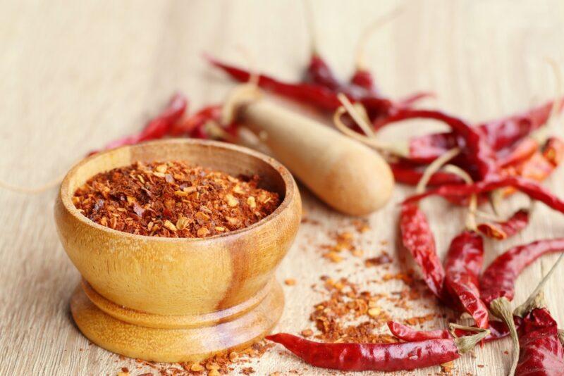 on a wooden surface is a wooden mortar and pestle with cayenne, around it are loose dried cayenne peppers and loose cayenne powder