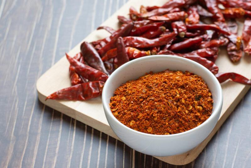 a closeup image of a white ceramic bowl full of cayenne powder with dried cayenne peppers behind it, all resting on top of a wooden chopping board