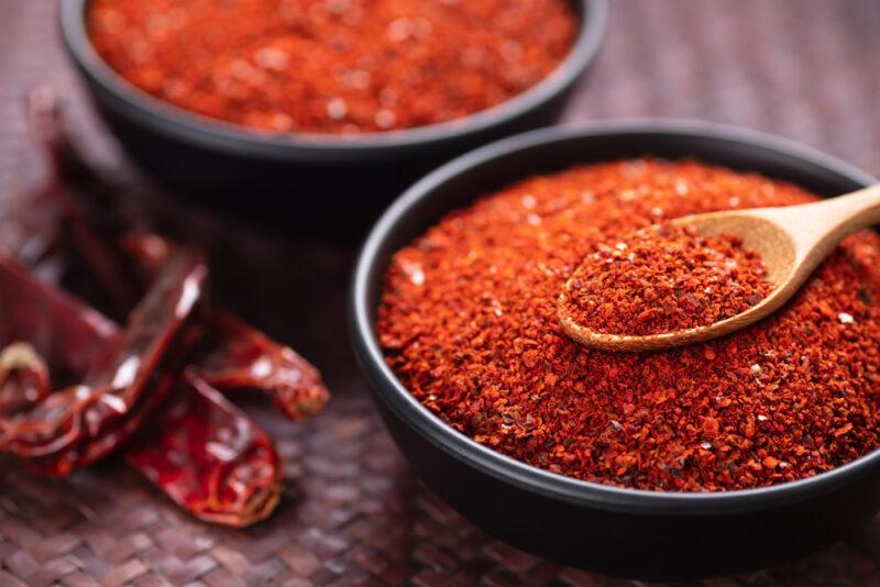 on a dark brown weaved table mat is a couple of black bowls full of cayenne pepper, one with a wooden spoon with cayenne pepper as well, beside the bowls are dried cayenne peppers