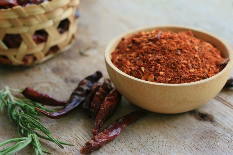 on a wooden surface is a wooden bowl full of cayenne powder, around it are loose dried cayenne, a sprig of fresh rosemary, and a small weaved basket full of dried cayenne peppers at the back