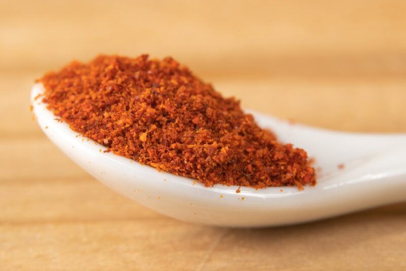 on a wooden surface is a closeup image of a white ceramic spoon with cayenne pepper