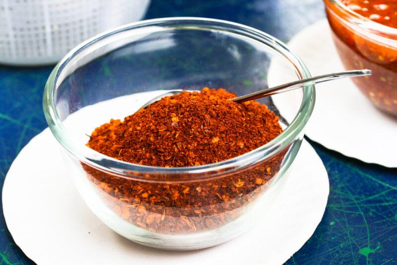 a clear glass bowl with cayenne pepper with a silver spoon shoved in it, resting on a white ceramic dish