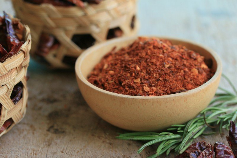 a small wooden bowl full of cayenne powder with rosemary herb and dried cayenne peppers beside it, with a couple of tiny native basket full of dried cayenne peppers.