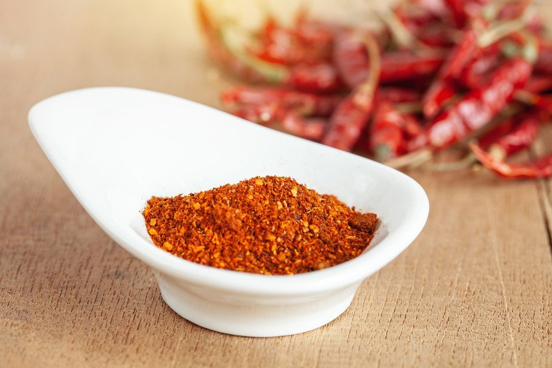 on a light brown surface is a white bowl with unique shape full of cayenne pepper, with dried cayenne peppers at the back