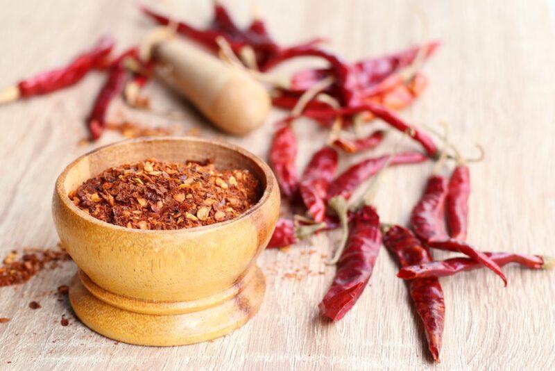 on a wooden surface is a wooden mortar and pestle full of ground dried cayenne, beside it are loose dried whole cayenne peppers