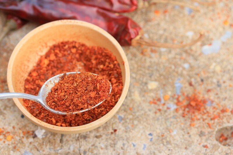 top view image of a wooden bowl with cayenne pepper, with a silver spoon in it, dried cayenne peppers at the back and loose cayenne powder around it