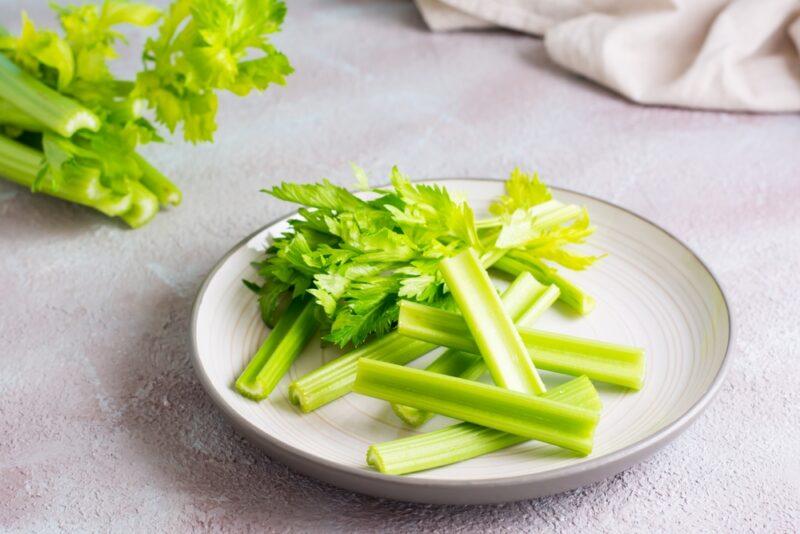 on a white surface is a white plate with celery stalks and leaves, behind it is a bunch of celery and white table cloth