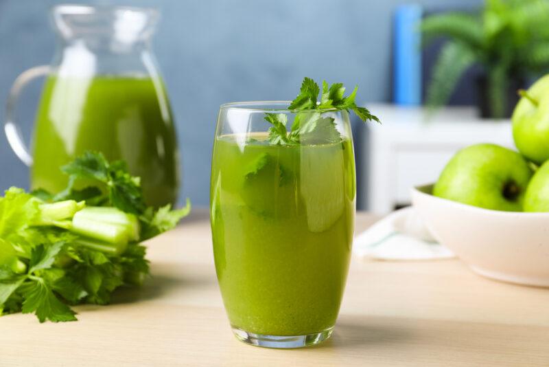 on a wooden surface is a tall glass of celery juice with fresh celery leaves on top, at the back is a bowl of green apples, a bunch of fresh celery, and a glass jug of celery juice