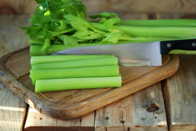 on a rustic looking wooden surface is a wooden chopping board with cut celery stalks and knife on it