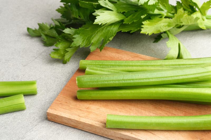 on a concrete surface is a wooden chopping board with celery sticks, with loose celery sticks beside it and celery leaves at the back