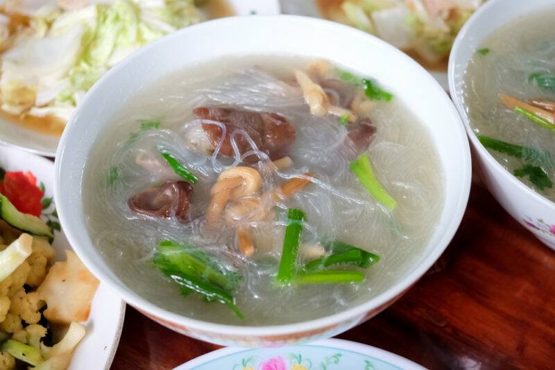 closeup image of a white bowl of chicken soup and cellophane noodles with toppings of chopped green onions and bits of meat