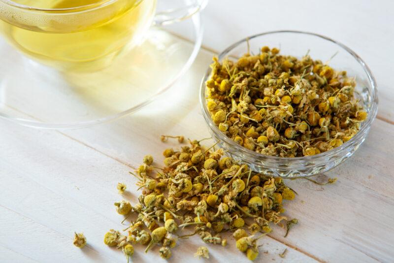 on a white wooden surface is a small glass dish with loose chamomile tea, beside it is a small pile of loose chamomile tea and a clear glass cup and saucer with tea