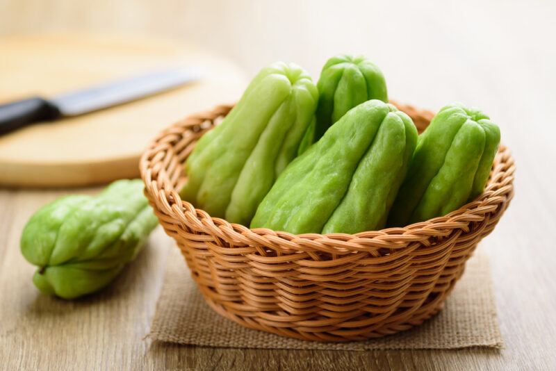 on a wooden surface is a weaved tray with four whole chayote with a chayote resting on a piece of burlap and chopping board with knife on it at the back