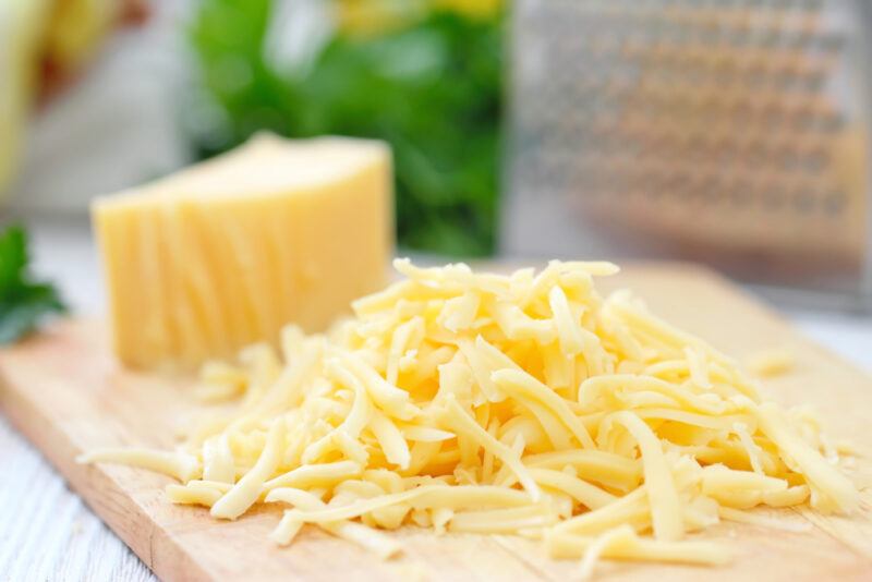 on a wooden chopping board is a big chunk and shredded cheese, at the back is a bunch of herbs beside a metal cheese grater