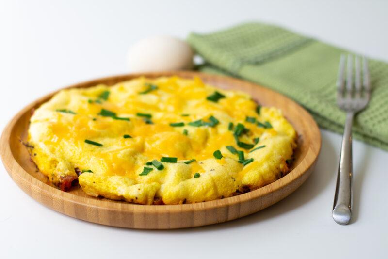 on a white surface is a wooden plate with cheese omelet with minced herbs on top, at the back is an egg and a green table napkin with silver fork on it