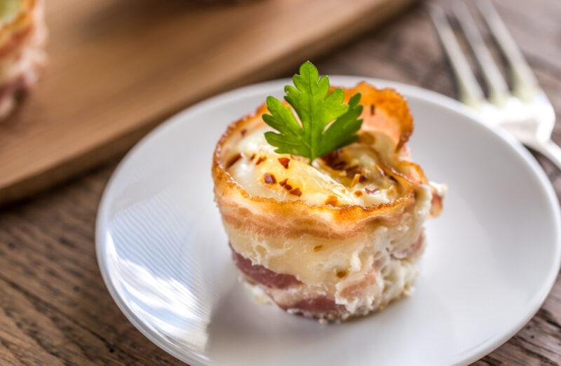 on a wooden surface is a white plate with a cheesy bacon egg cups with a sprig of herb on top, beside it is a silver fork, at the back is a silk gold table napkin