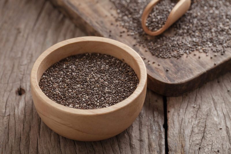 on a rustic wooden surface is a wooden bowl with chia seeds, at the back is a wooden board with chia seeds and wooden scoop on it