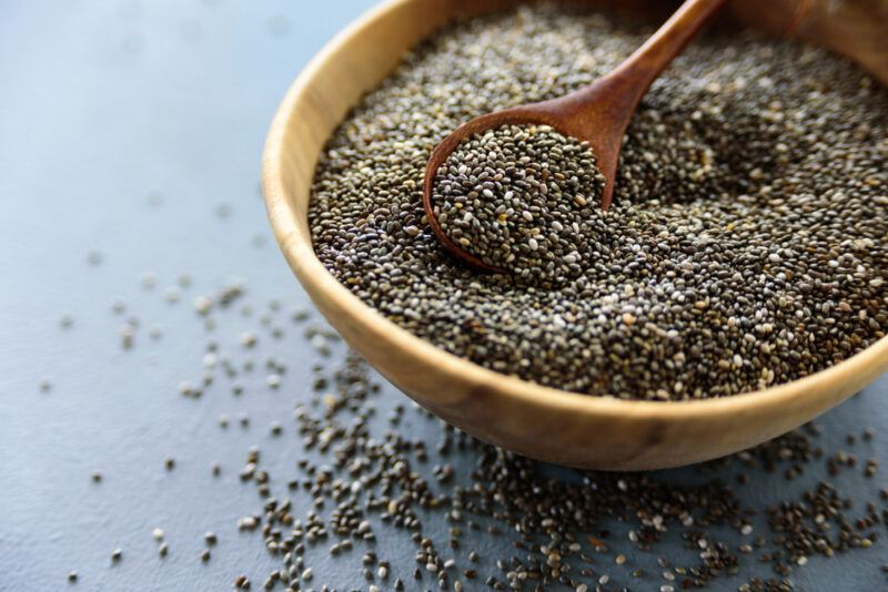 on a dark colored surface is a wooden bowl full of chia seeds with a wooden spoon shoved in it, around it are loose chia seeds