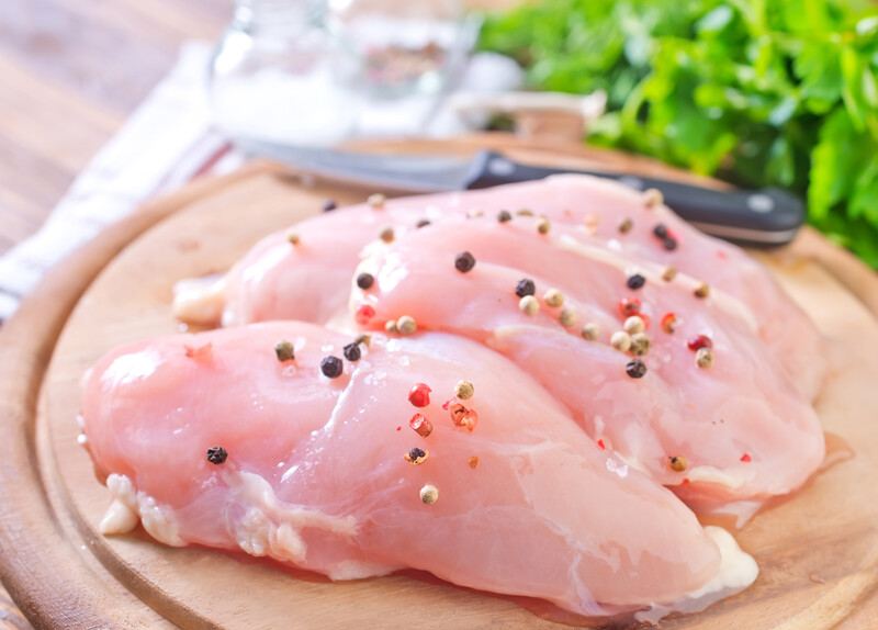 closeup image of chicken breast on a wooden round chopping board with herbs and spices on it and knife at the back