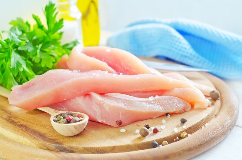 chicken breast fillet on a wooden chopping board with spices and herbs, with a jar of olive oil and blue table napkin at the back