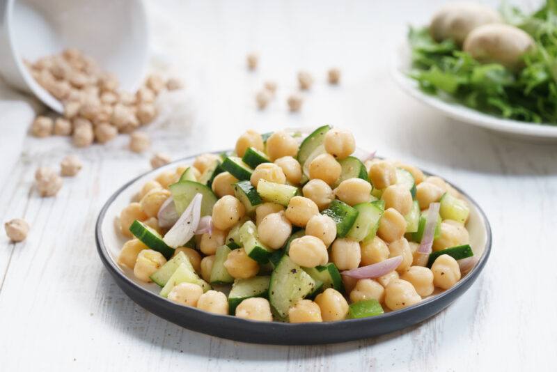 on a white surface is a black and white dish with chickpea salad, around it are loose chickpeas, a small ceramic jar and a white plate of greens