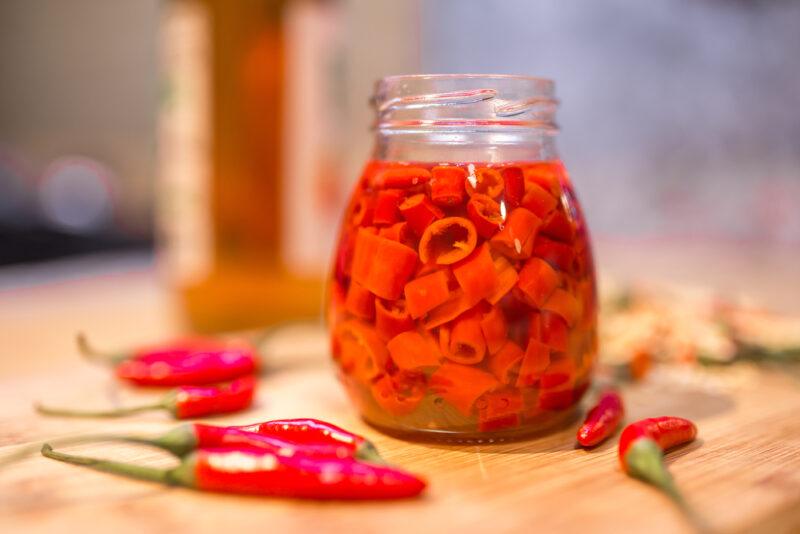 on a wooden chopping board is a jar of chopped pickled chili peppers, around it are fresh red chili peppers and a bottle at the back 