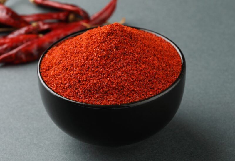 on a dark surface is a black wooden bowl full of chili powder with dried chili peppers behind it