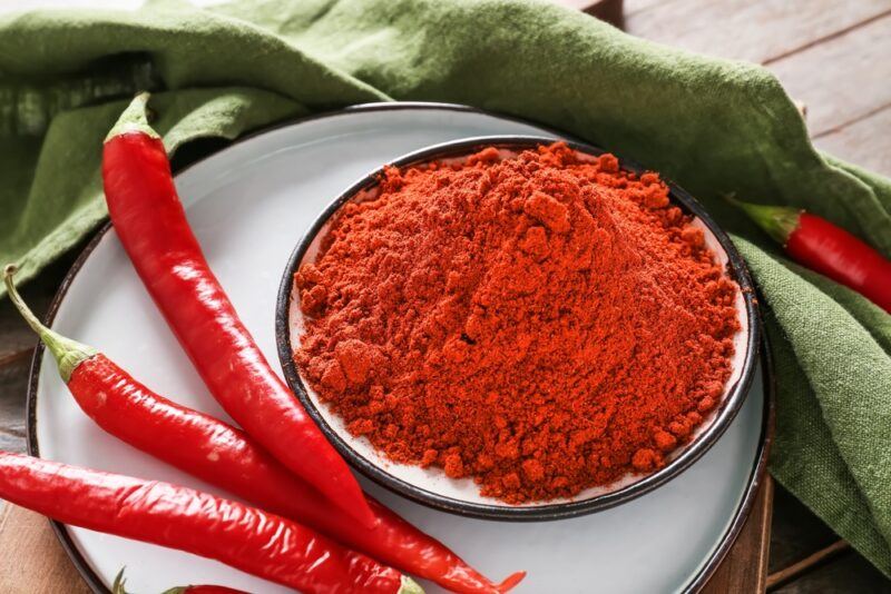 on a wooden surface is a closeup image of a bowl full of chili powder resting on a white dish with fresh chilis beside it, a green colored table napkin is draped behind it