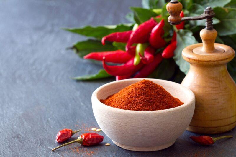 on a dark wooden surface is a white bowl full of chili powder with a brown pepper grinder and fresh chilis at the back, with a couple of loose dried chilis beside it