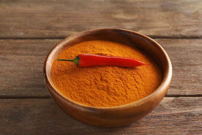 on a rustic looking wooden surface is a wooden bowl with chili powder with one fresh red chili on top