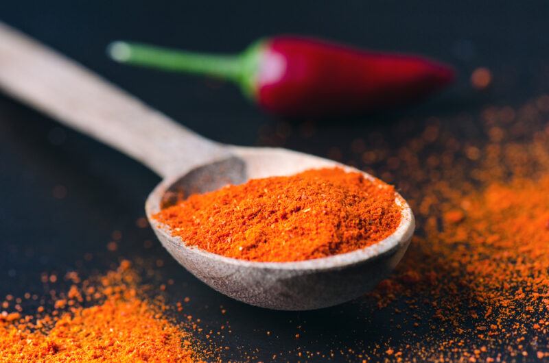 on a dark black surface is a wooden spoon with chili powder with loose chili powder and a fresh red chili at the back