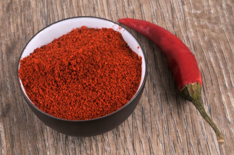 on a wooden surface is a black and white ceramic bowl of chili powder with a fresh red chili beside it