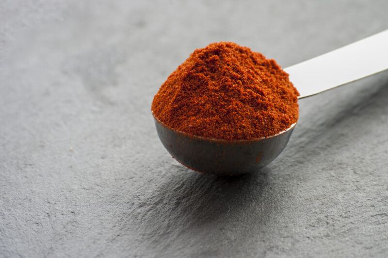 on a black concrete surface is a metal measuring spoon full of chili powder