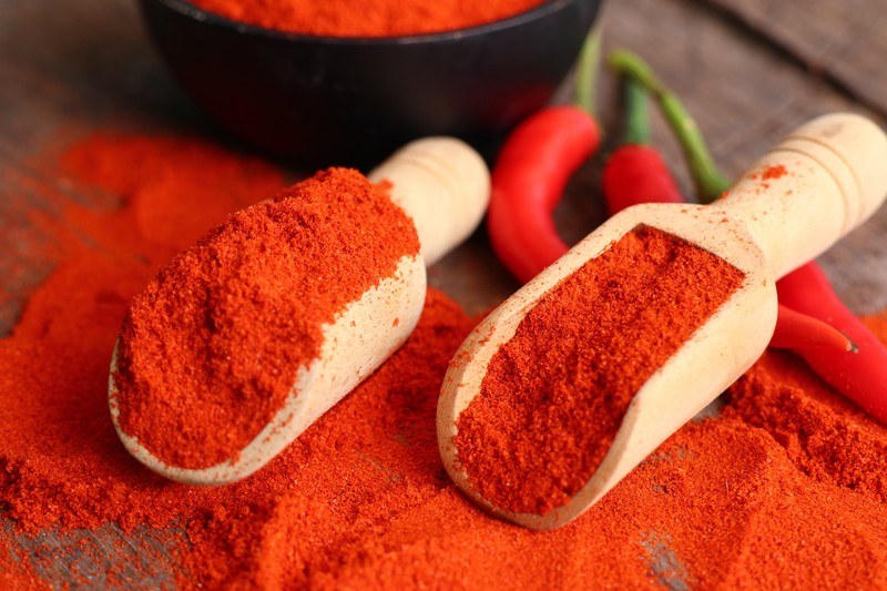 closeup image of two wooden scoops full of chili powder resting on top of a wooden surface with spilled chili powder and fresh chilis underneath it