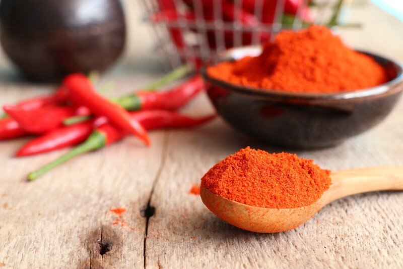 closeup image of a wooden surface with a wooden spoon full of chili powder with a black bowl full of chili powder and fresh chili at the back