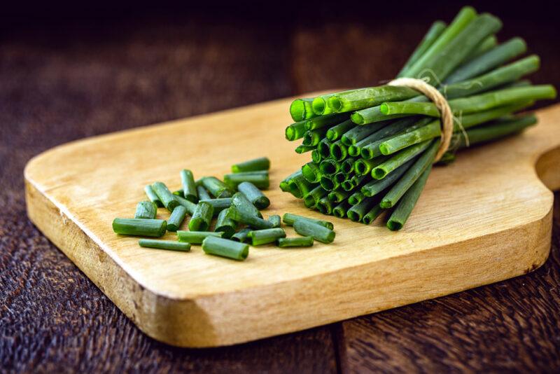 on a dark brown wooden surface is a light brown chopping board with a partially cut bunch of fresh chives