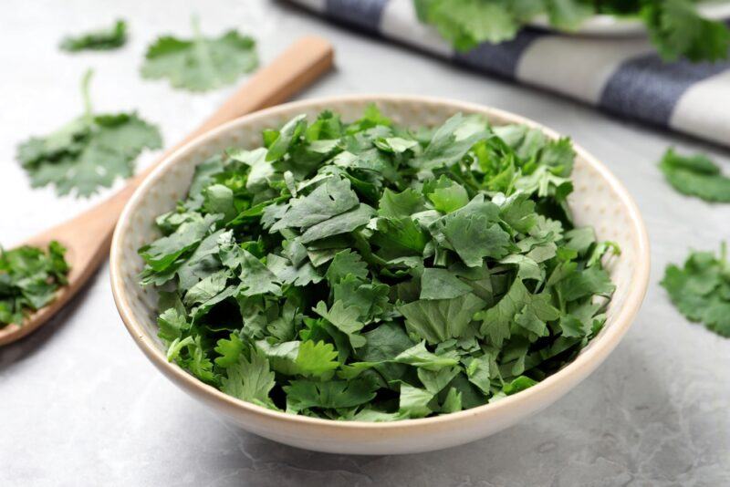 closeup image of a light brown ceramic bowl full of chopped cilantro, with a wooden spoon beside it, and white and blue checkered table napkin at the back,  with loose cilantro around