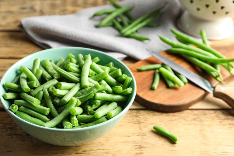 on a wooden surface is a bowl of chopped green beans at the back is a grey table napkin and wooden chopping board with chopped and whole green beans with a knife on it