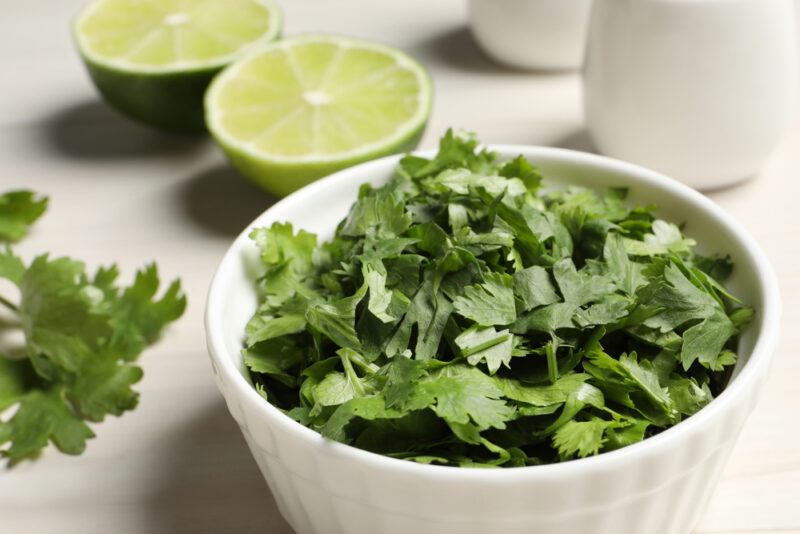 on a white surface is a white bowl full of fresh chopped cilantro, with loose chopped cilantro around it, behind is a halved lime and a couple of white small ceramic jars