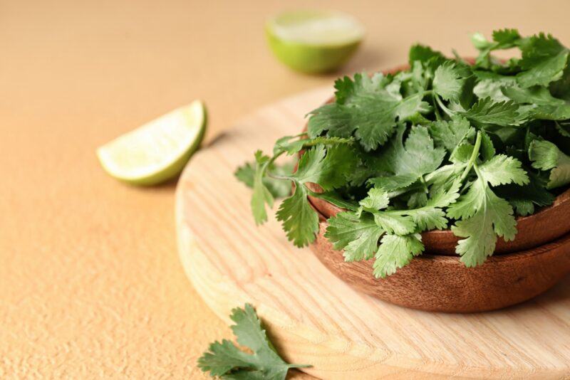 on a light brown surface is a light brown wooden chopping board with a dark brown wooden bowl full of fresh cilantro, at the back is a couple of lime wedges