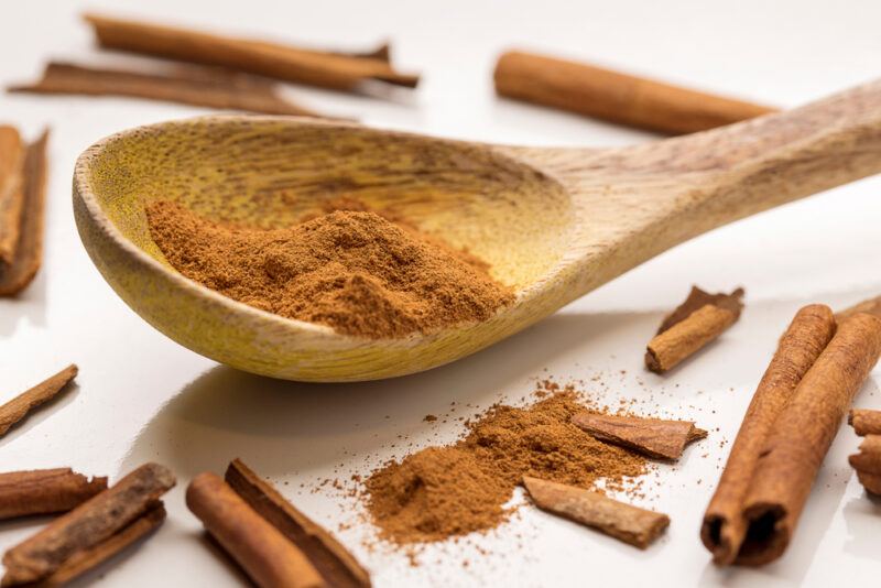 on a white surface is a wooden ladle with ground cinnamon, with cinnamon sticks and loose cinnamon powder around it