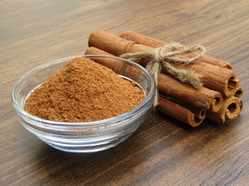 on a wooden surface is a clear glass bowl of ground cinnamon with a bundle of cinnamon sticks behind it