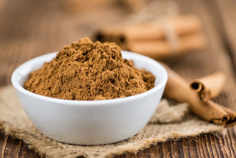 on a wooden surface is a white bowl full of ground cinnamon resting on a burlap sack with cinnamon sticks at the back