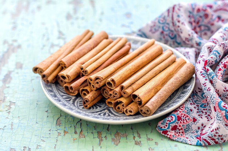 on a rustic green wooden surface is a printed white and blue plate with cinnamon sticks, at the back of the plate is a bluish and reddish floral-printed cloth