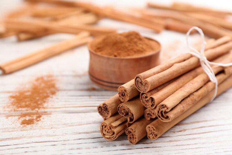 on a faded white wooden surface is a bundle of cinnamon sticks tied together with a white string, behind it is a small bowl of ground cinnamon, loose cinnamon powder and sticks