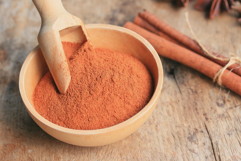 a wooden bowl full of cinnamon powder with a wooden scoop, resting on an aged wooden surface with a couple of cinnamon sticks at the back 