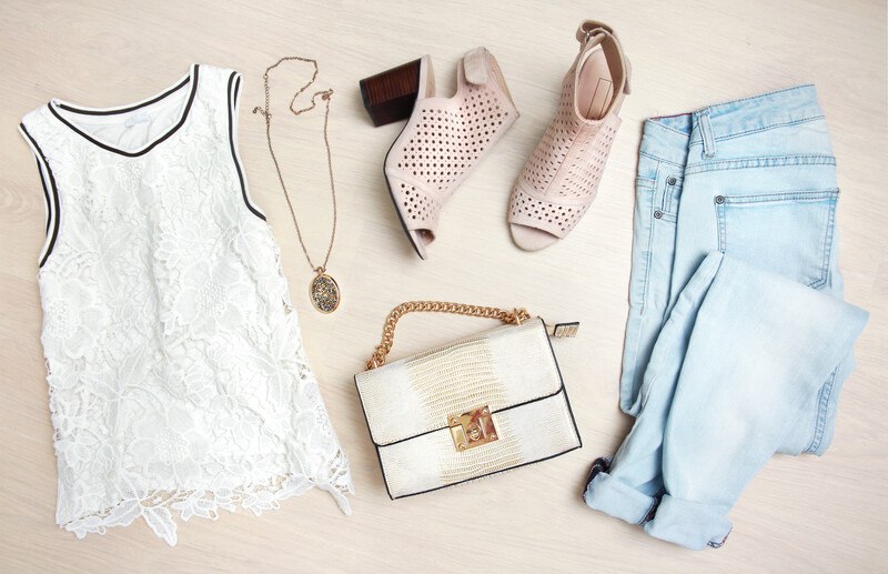 clothes and accessories laid out on a light wood background white lace sleeveless top with black outline, pendant necklace, high heel shoes, medium sized clutch, and light colored blue jeans