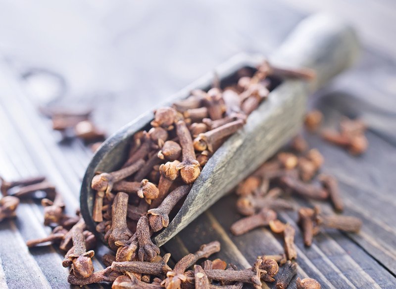 a closeup image of an aged wooden scoop full of cloves resting on a wooden surface with some of the cloves spilling all around it