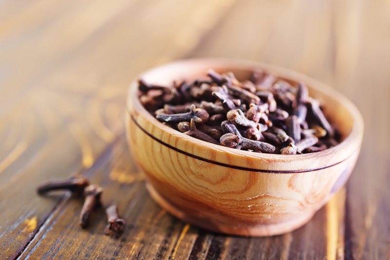 on a wooden surface is a closeup image of a wooden bowl full of cloves with 3 cloves beside it